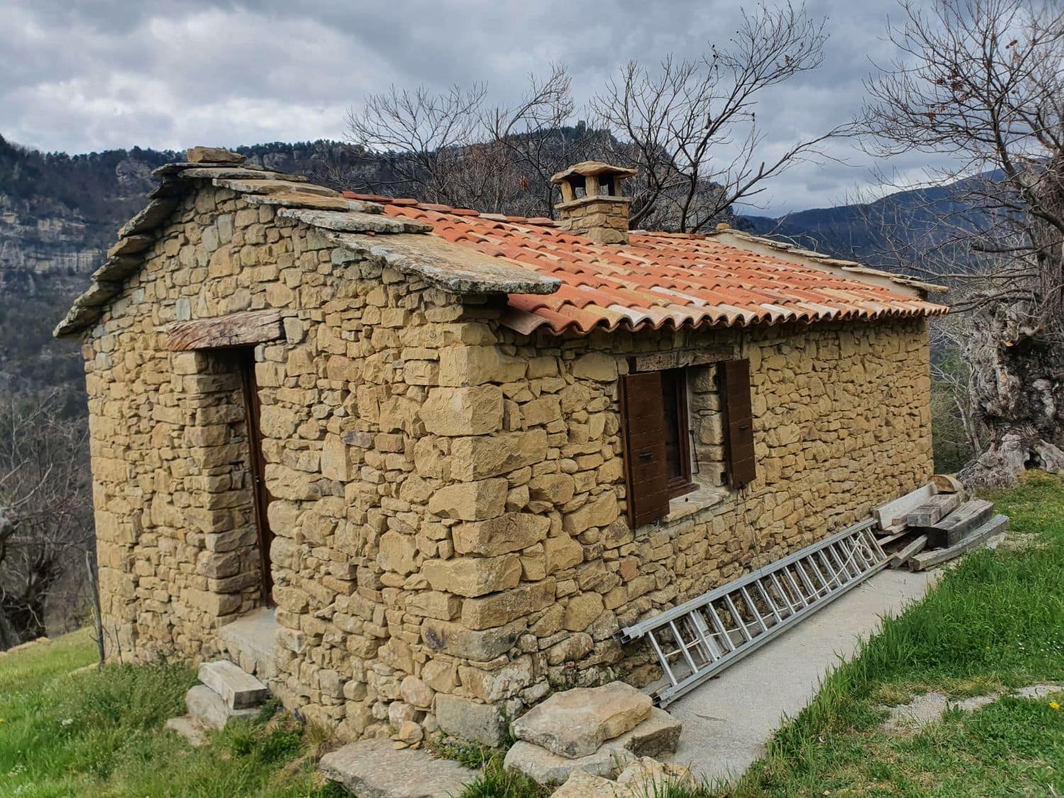 Stone hut, tiled roof with stone slab verges, facing the mountain.