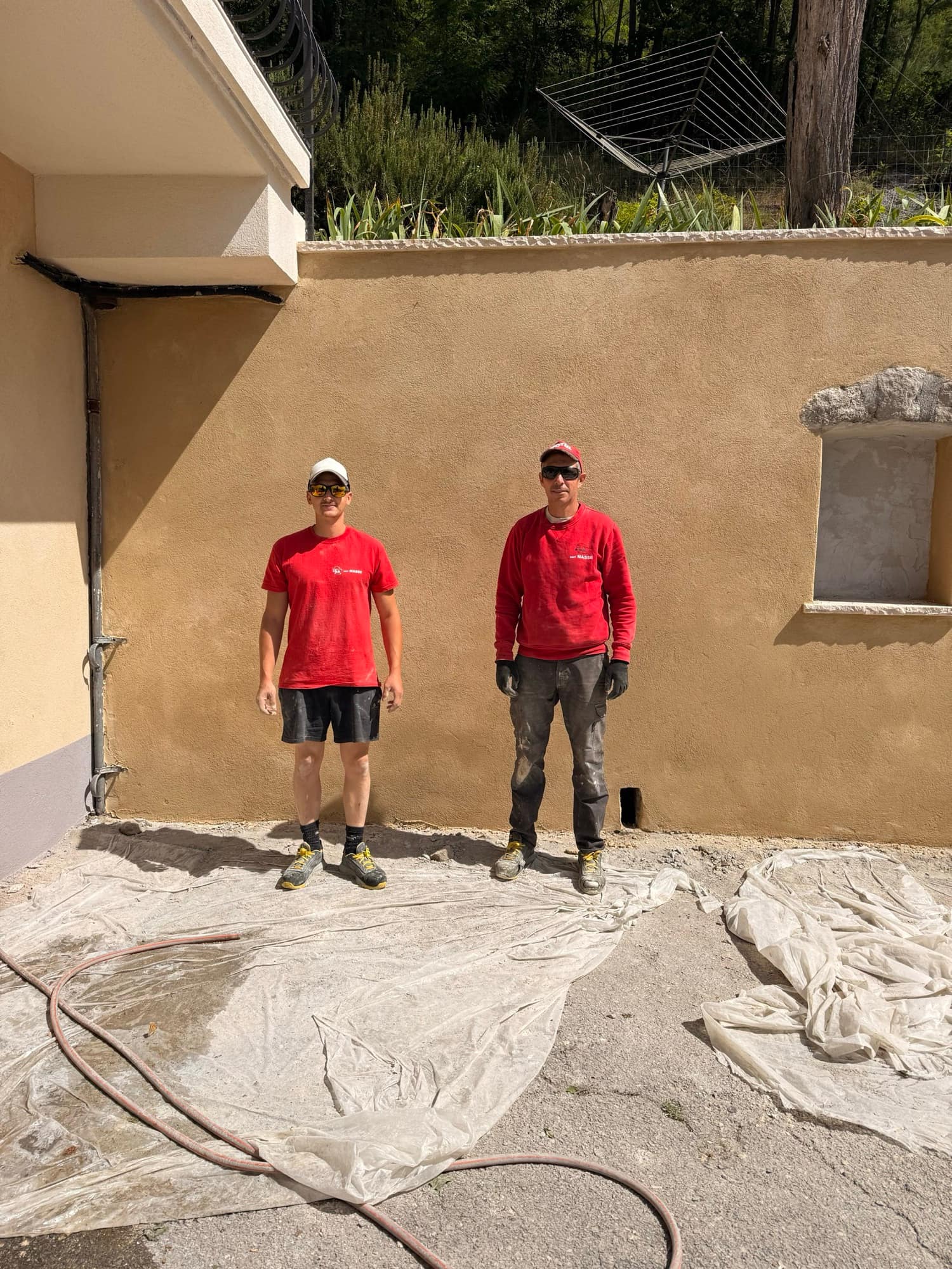 Olivier and Franck Masse in front of a freshly rendered sand-coloured façade, in red work clothes.