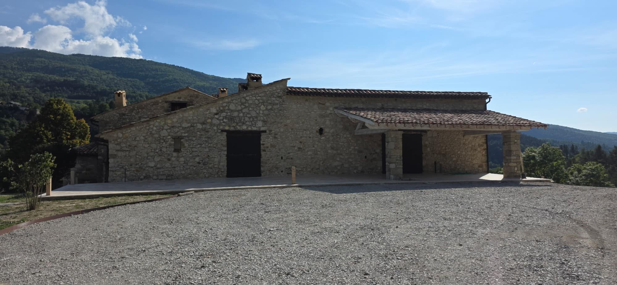Long stone building and its porch on stone piers, tiled roof redone, gravelled yard facing the mountains.