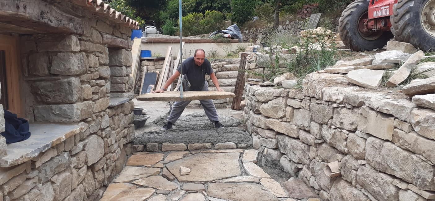 Mason carrying a stone slab to lay it, between two dry-stone walls.