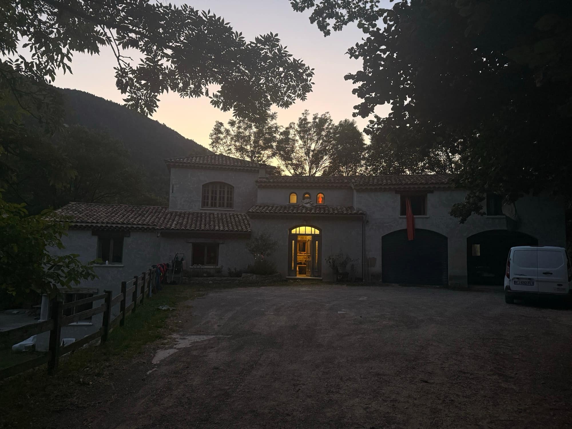 Stone house built by SARL MASSE, lit from within at dusk, wooded mountain behind.